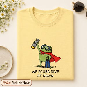 May include: A yellow haze t-shirt featuring a green frog wearing a red cape, blue scuba goggles, and blue flippers. The frog holds a grey scuba tank. Below the illustration, the text reads: WE SCUBA DIVE AT DAWN in black, sans-serif capital letters.