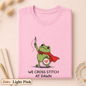 May include: A light pink t-shirt featuring a green frog wearing a red cape, holding a needle with red thread and an embroidery hoop with a heart design. Below the frog, the text reads: WE CROSS STITCH AT DAWN. The shirt has a crew neckline.