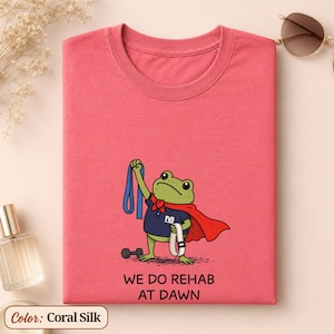 Op de afbeelding: Een koraalkleurig zijden T-shirt met een afbeelding van een groene kikker in een rode cape en blauw shirt, die een blauwe weerstandsband en een witte riem vasthoudt. De tekst WE DO REHAB AT DAWN staat in zwarte letters onder de kikker.
