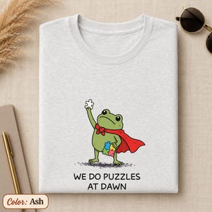 Könnte beinhalten: Ein aschefarbenes T-Shirt mit einer Grafik eines grünen Frosches, der einen roten Umhang trägt. Der Frosch hält ein weißes Puzzleteil in der einen und ein buntes Puzzleteil in der anderen Hand. Darunter steht der Text: WE DO PUZZLES AT DAWN.