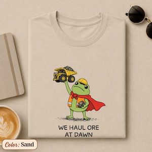 Könnte beinhalten: Ein sandfarbenes T-Shirt mit einer grünen Frosch-Illustration, der einen gelben Bauhelm und ein rotes Cape trägt und einen mit Steinen beladenen gelben Muldenkipper hochhält. Darunter steht der Text WE HAUL ORE AT DAWN in schwarzen Buchstaben.