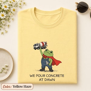 May include: A yellow haze t-shirt featuring a green frog wearing blue overalls and a red cape, holding a cement mixer truck above its head. The text below the frog reads WE POUR CONCRETE AT DAWN in black capital letters.