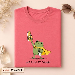 May include: A coral-coloured t-shirt featuring a graphic of a green frog wearing a yellow cape, holding a yellow trainer in one hand and a water bottle in the other. Below the frog, the text reads WE RUN AT DAWN in black capital letters.