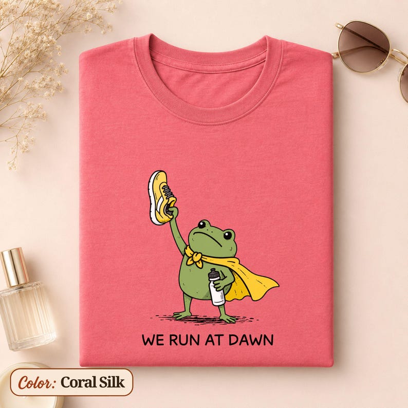 May include: A coral-coloured t-shirt featuring a graphic of a green frog wearing a yellow cape, holding a yellow trainer in one hand and a water bottle in the other. Below the frog, the text reads WE RUN AT DAWN in black capital letters.