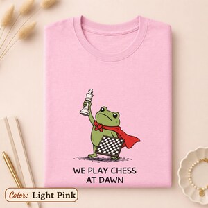 May include: A light pink t-shirt featuring a green frog wearing a red cape, holding a white chess king piece in one hand and a black and white checkered board in the other, with the text "WE PLAY CHESS AT DAWN" printed below the illustration.