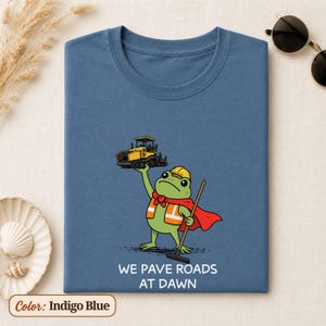 May include: An indigo blue t-shirt featuring a graphic of a green frog wearing a yellow hard hat, an orange safety vest, and a red cape. The frog holds a yellow road paver above its head and a rake in its hand. Text reads: WE PAVE ROADS AT DAWN.