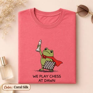 May include: A coral silk t-shirt featuring a green frog wearing a red cape, holding a white chess piece, and carrying a chessboard. Below the illustration, the text reads WE PLAY CHESS AT DAWN in black capital letters. The shirt has a crew neckline.
