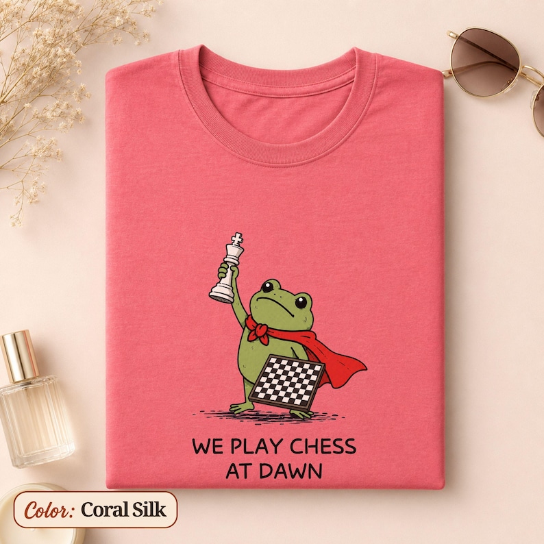May include: A coral silk t-shirt featuring a green frog wearing a red cape, holding a white chess piece, and carrying a chessboard. Below the illustration, the text reads WE PLAY CHESS AT DAWN in black capital letters. The shirt has a crew neckline.