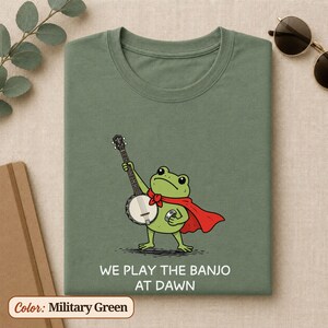 K&ouml;nnte beinhalten: Ein milit&auml;rgr&uuml;nes T-Shirt mit einer Grafik eines gr&uuml;nen Frosches, der einen roten Umhang tr&auml;gt und Banjo spielt. Der Frosch h&auml;lt einen kleinen silbernen Gegenstand in der anderen Hand. Unter der Abbildung steht der Text: WE PLAY THE BANJO AT DAWN.