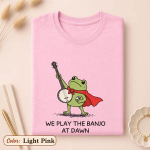 K&ouml;nnte beinhalten: Ein hellrosa T-Shirt mit einer gr&uuml;nen Frosch-Illustration, der einen roten Umhang tr&auml;gt und Banjo spielt. Der Text unter dem Frosch lautet "WE PLAY THE BANJO AT DAWN". Das T-Shirt hat einen Rundhalsausschnitt und kurze &Auml;rmel, flach zusammengelegt.