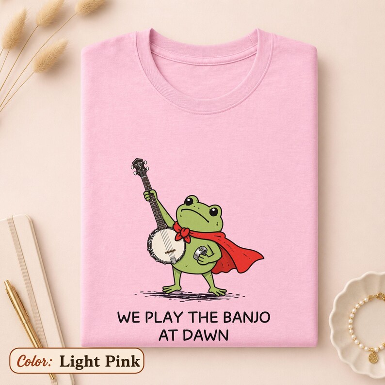 K&ouml;nnte beinhalten: Ein hellrosa T-Shirt mit einer gr&uuml;nen Frosch-Illustration, der einen roten Umhang tr&auml;gt und Banjo spielt. Der Text unter dem Frosch lautet "WE PLAY THE BANJO AT DAWN". Das T-Shirt hat einen Rundhalsausschnitt und kurze &Auml;rmel, flach zusammengelegt.