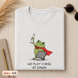 May include: An ash-colored t-shirt featuring a graphic of a green frog wearing a red cape, holding a white chess piece above its head, and carrying a chessboard. Below the illustration, the text reads: WE PLAY CHESS AT DAWN in black letters.