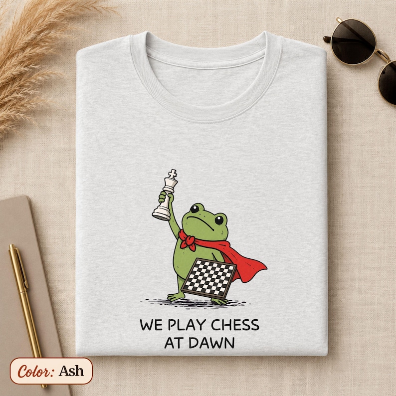 May include: An ash-colored t-shirt featuring a graphic of a green frog wearing a red cape, holding a white chess piece above its head, and carrying a chessboard. Below the illustration, the text reads: WE PLAY CHESS AT DAWN in black letters.