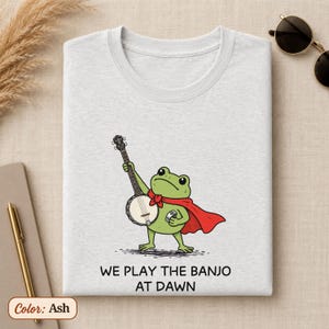 K&ouml;nnte beinhalten: Ein aschefarbenes T-Shirt mit einer Grafik eines gr&uuml;nen Frosches, der einen roten Umhang tr&auml;gt und Banjo spielt. Unter der Abbildung steht der Text: WE PLAY THE BANJO AT DAWN. Das T-Shirt hat einen Rundhalsausschnitt und kurze &Auml;rmel.
