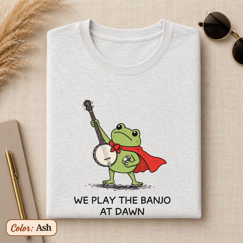 K&ouml;nnte beinhalten: Ein aschefarbenes T-Shirt mit einer Grafik eines gr&uuml;nen Frosches, der einen roten Umhang tr&auml;gt und Banjo spielt. Unter der Abbildung steht der Text: WE PLAY THE BANJO AT DAWN. Das T-Shirt hat einen Rundhalsausschnitt und kurze &Auml;rmel.