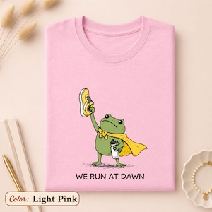 May include: A light pink t-shirt featuring a graphic of a green frog wearing a yellow cape, holding a yellow running shoe in one hand and a water bottle in the other. Below the frog, the text reads WE RUN AT DAWN in black capital letters.
