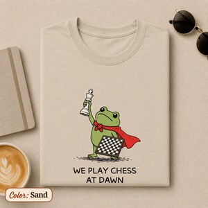 May include: A sand-colored t-shirt featuring a graphic of a green frog wearing a red cape, holding a white chess king piece and a chessboard. Below the illustration, the text reads: WE PLAY CHESS AT DAWN. The design is centered on the chest.