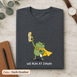 May include: A dark heather grey t-shirt featuring a green frog wearing a yellow cape, holding a yellow running shoe in one hand and a white water bottle in the other, with the white text "WE RUN AT DAWN" printed below the graphic illustration.