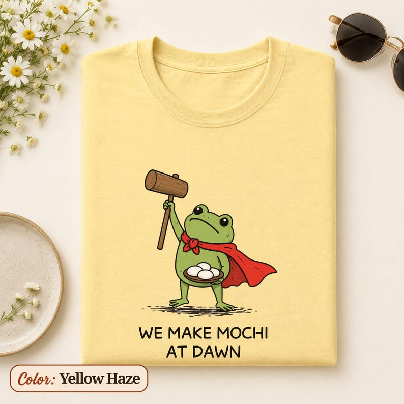 K&ouml;nnte beinhalten: Ein hellgelbes T-Shirt mit einer gr&uuml;nen Frosch-Illustration, der einen roten Umhang tr&auml;gt, einen Holzhammer h&auml;lt und ein Tablett mit wei&szlig;en Mochi tr&auml;gt. Unter dem Bild steht der Text: WE MAKE MOCHI AT DAWN. Mit Rundhalsausschnitt.