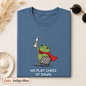 May include: An indigo blue t-shirt featuring a graphic of a green frog wearing a red cape, holding a white chess king piece in one hand and a chessboard in the other. Below the illustration, the text reads: WE PLAY CHESS AT DAWN in white letters.