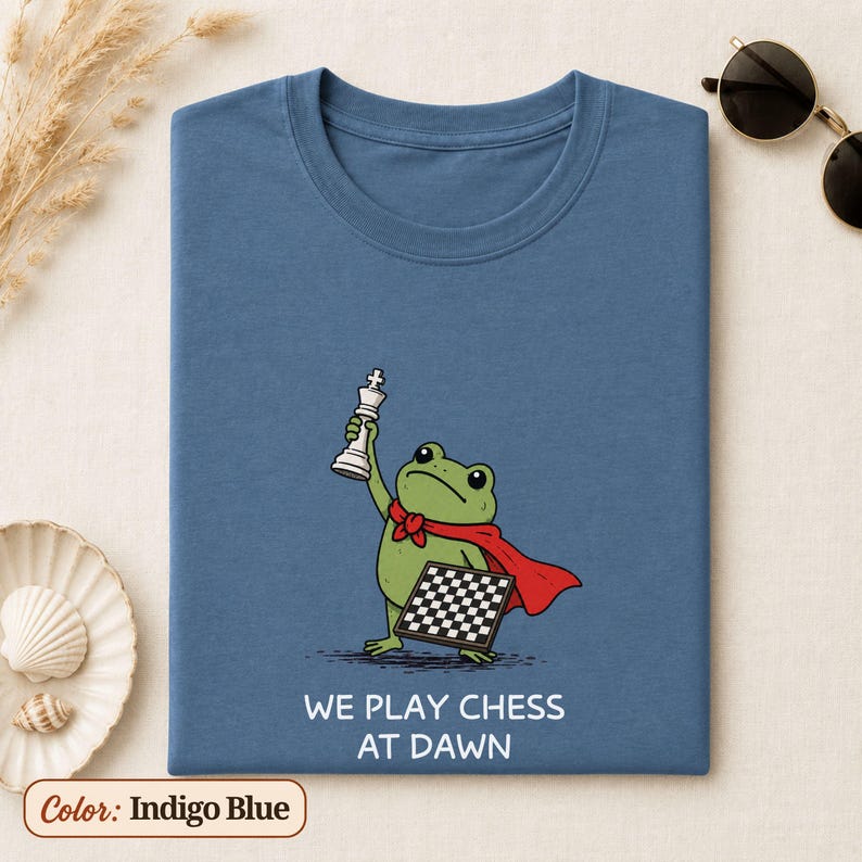 May include: An indigo blue t-shirt featuring a graphic of a green frog wearing a red cape, holding a white chess king piece in one hand and a chessboard in the other. Below the illustration, the text reads: WE PLAY CHESS AT DAWN in white letters.
