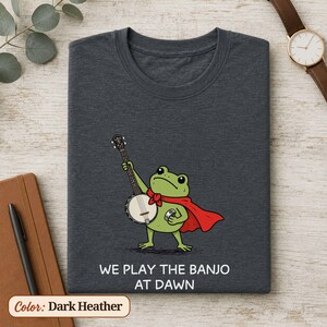 K&ouml;nnte beinhalten: Ein dunkelgrau meliertes T-Shirt mit einer Grafik eines gr&uuml;nen Frosches, der einen roten Umhang tr&auml;gt und Banjo spielt. Unter der Abbildung steht der Text "WE PLAY THE BANJO AT DAWN" in wei&szlig;en Gro&szlig;buchstaben. Das T-Shirt hat einen Rundhalsausschnitt.