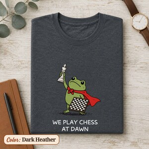 May include: A dark heather gray t-shirt featuring a green frog wearing a red cape, holding a white chess king piece in one hand and a chessboard in the other. Below the graphic, the text reads WE PLAY CHESS AT DAWN in white capital letters.