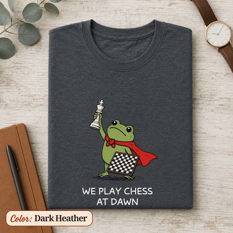May include: A dark heather gray t-shirt featuring a green frog wearing a red cape, holding a white chess king piece in one hand and a chessboard in the other. Below the graphic, the text reads WE PLAY CHESS AT DAWN in white capital letters.