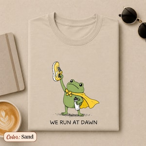 May include: A sand-coloured t-shirt featuring a graphic of a green frog wearing a yellow cape, holding a yellow running shoe in one hand and a water bottle in the other. Below the illustration, the text reads WE RUN AT DAWN in black letters.