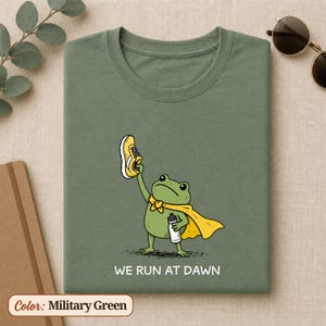 May include: A military green t-shirt featuring a graphic of a green frog wearing a yellow cape, holding a yellow trainer in one hand and a white water bottle in the other, with the text "WE RUN AT DAWN" printed in white below the illustration.
