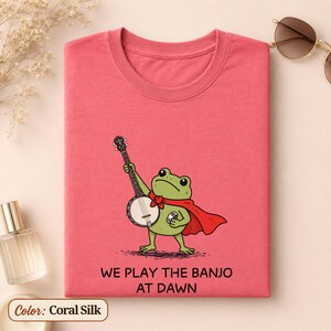 K&ouml;nnte beinhalten: Ein korallenseidenfarbenes T-Shirt mit einer gr&uuml;nen Frosch-Illustration, der einen roten Umhang tr&auml;gt und Banjo spielt. Unter dem Frosch steht der Text: WE PLAY THE BANJO AT DAWN. Das T-Shirt hat einen Rundhalsausschnitt und kurze &Auml;rmel.