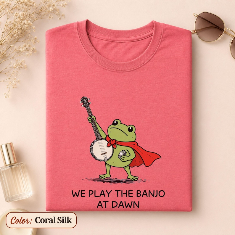 K&ouml;nnte beinhalten: Ein korallenseidenfarbenes T-Shirt mit einer gr&uuml;nen Frosch-Illustration, der einen roten Umhang tr&auml;gt und Banjo spielt. Unter dem Frosch steht der Text: WE PLAY THE BANJO AT DAWN. Das T-Shirt hat einen Rundhalsausschnitt und kurze &Auml;rmel.