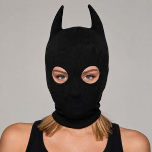 May include: A solid black knit balaclava featuring two prominent, pointed horn-like structures at the top. The headgear is designed with two circular eye openings, providing a full-face covering that extends down to the neck with a ribbed hem.