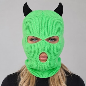 May include: A bright neon green knitted balaclava featuring two prominent black devil horns attached to the top. The headgear is constructed with a ribbed knit texture and includes two eye openings and a single mouth opening for full facial coverage.