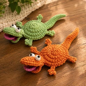 May include: Two handmade crochet lizards in the image. One is green, the other is orange. Both have white eyes with black pupils and pink tongues sticking out. The lizards are lying on a wooden surface.