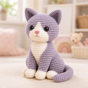 May include: A handmade crochet cat in a seated position. The cat is primarily lavender with white accents on the chest, paws, and face. It has large black eyes, a pink nose, and a long tail. The cat is sitting on a cream-colored surface.
