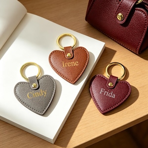 May include: Three heart-shaped keychains with gold-tone metal rings and studs. The gray keychain is embossed with the name Cindy, the brown one with Irene, and the burgundy one with Frida. Each keychain features a textured surface finish.