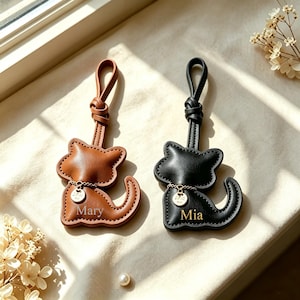 Può includere: Due ciondoli per borsa a forma di gatto in pelle, uno marrone e uno nero. Ogni ciondolo presenta un cinturino ad anello, una piccola catenina dorata con un ciondolo circolare e un nome inciso sul corpo: Mary sul gatto marrone e Mia su quello nero.