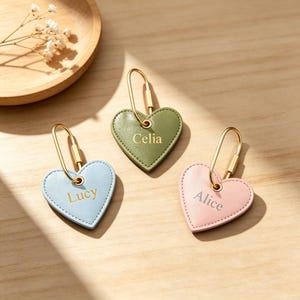 May include: Three heart-shaped keychains in light blue, olive green, and soft pink. Each keychain features a gold-toned metal loop and is engraved with a name: Lucy on the blue, Celia on the green, and Alice on the pink heart-shaped tag.