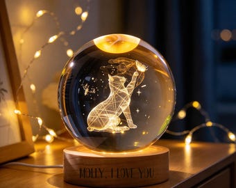 May include: A clear glass sphere lamp featuring an internal 3D laser-etched cat reaching for a butterfly. The sphere rests on a circular wooden base with the engraved text "MOLLY, I LOVE YOU." The lamp emits a warm, golden glow.