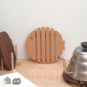 May include: A light-coloured wooden trivet shaped like a fish with vertical slats, positioned upright. To the left, a darker wooden fish-shaped trivet rests on a stand. To the right, a stainless steel gooseneck kettle sits on a dark wooden base.