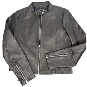 May include: A black leather jacket featuring a stand-up collar with a snap button closure, a full-length silver-tone front zipper, and long sleeves with zippered cuffs. The jacket includes vertical seam detailing and a label at the inner neckline.