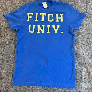 May include: A short-sleeved blue t-shirt featuring the text FITCH UNIV. printed in yellow across the chest. The garment has a crew neckline and a standard fit, with a white brand label visible inside the collar at the back of the neck.