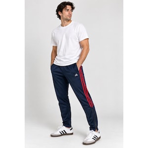 Puede incluir: Una camiseta blanca lisa de manga corta combinada con un pantalón de chándal azul marino con tres rayas verticales rojas en los laterales, un pequeño logotipo blanco en el muslo y cremalleras en los bajos, junto con zapatillas blancas.