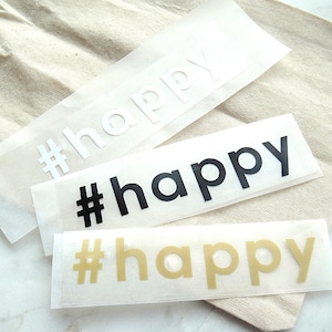 Könnte beinhalten: Drei rechteckige Vinyl-Aufkleber mit dem Hashtag-Symbol gefolgt von dem Wort happy in Kleinbuchstaben. Der oberste Aufkleber ist weiß, der mittlere ist schwarz und der untere ist in einer metallischen Goldfarbe auf einer transparenten Trägerfolie.