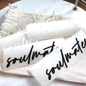 Könnte beinhalten: Mehrere selbstklebende Vinyl-Aufkleber mit dem Wort soulmates in einer kursiven Schriftart. Die Sammlung umfasst schwarze, weiße und transparente Schriftzüge, die jeweils auf einer rechteckigen Trägerfolie auf einem beigen Stoffbeutel liegen.