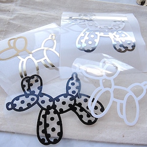 Könnte beinhalten: Vier Ballonhund-Aufkleber auf transparenter Transferfolie. Ein Aufkleber ist goldfarben, einer ist silberfarben mit Punkten, einer ist schwarz mit Punkten und einer ist weiß. Diese Klebemotive zeigen die klassische Ballon-Tierform in verschiedenen Ausführungen.