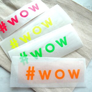 Könnte beinhalten: Vier rechteckige Klebetransferfolien mit dem Text #WOW in Neonpink, Neongelb, Neongrün und leuchtendem Orange. Jeder Aufkleber ist auf halbtransparentem Trägerpapier gedruckt und zeigt eine kräftige, serifenlose Schrift in leuchtenden Farben.
