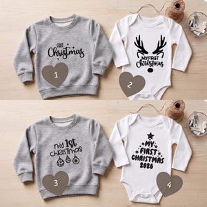 Könnte beinhalten: Zwei graue Langarm-Sweatshirts und zwei weiße Langarm-Bodys mit schwarzem Text und Grafiken. Das erste Sweatshirt zeigt First Christmas, das zweite my 1st Christmas mit Ornamenten. Die Bodys zeigen ein Rentiergeweih und einen Weihnachtsbaum mit dem Text My First Christmas 2026.