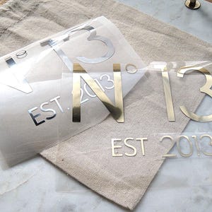 Könnte beinhalten: Zwei transparente Vinyl-Aufkleber mit metallisch goldener Schrift. Die Designs zeigen den Text N° 13 und EST. 2013 in einer klaren serifenlosen Schriftart. Diese Transferaufkleber liegen auf einem hellbeigen Stoffbeutel mit sichtbarer Webstruktur.